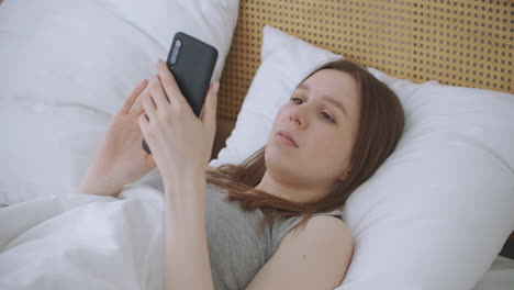 Woman-in-morning-sitting-in-bedroom-leaning-against-the-soft-leather-back-of-bed.-Write-messages-with-your-smartphone-use-apps-to-information